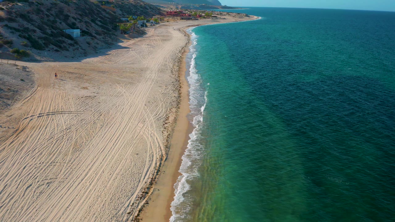 Drone-like aerial frame over El Sargento La Ventana, Mexico, with the surf line centered more vertically, pale raked-looking sand to the left, blue-green water to the right, and no visible people.