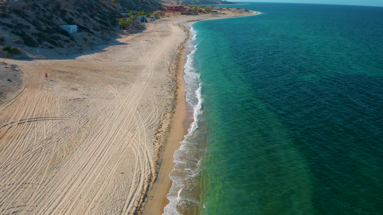 Aerial coastal view in El Sargento La Ventana, Mexico, with more open water filling the right side, a curving sandy shoreline at upper frame, small wave foam, and no visible people.