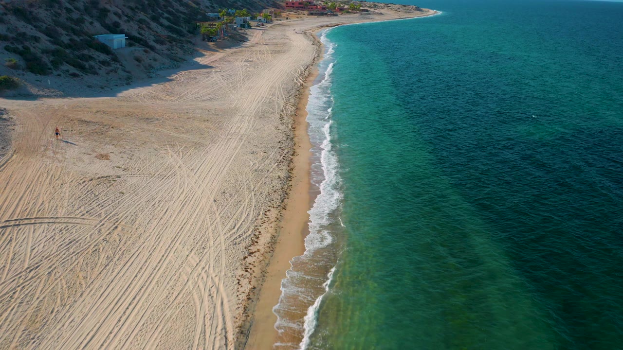 Mid-clip aerial frame over El Sargento La Ventana, Mexico, showing hillside shadow across the upper beach, a narrow wet sand strip, foamy wave edge, green-blue shallows, and no visible people.