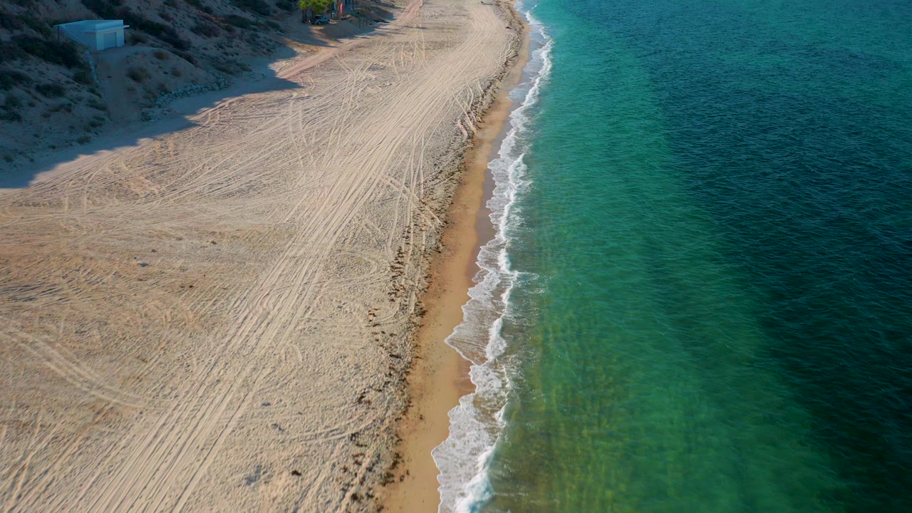 High aerial view in El Sargento La Ventana, Mexico, showing tracked tan sand, a narrow surf line, turquoise water, and no visible people at the start of the clip.