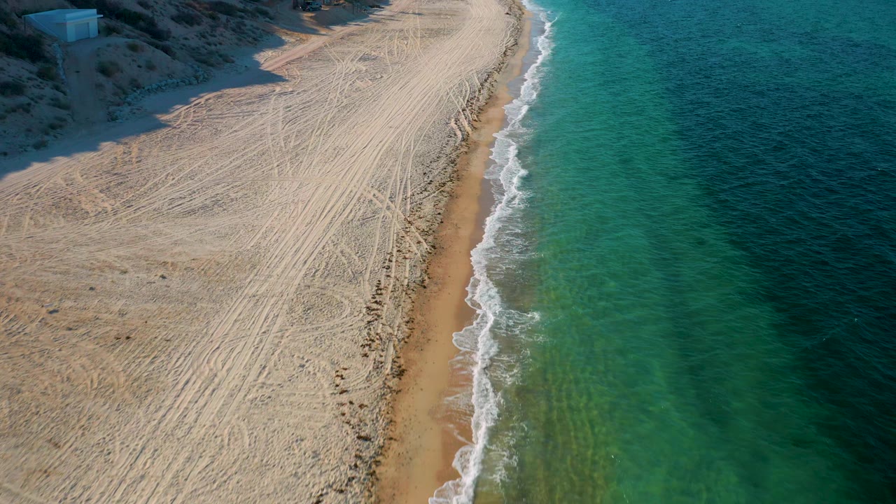 Aerial frame in El Sargento La Ventana, Mexico, showing dense tire tracks across pale sand, a thin amber wet-sand strip, clear blue water, and no visible people.