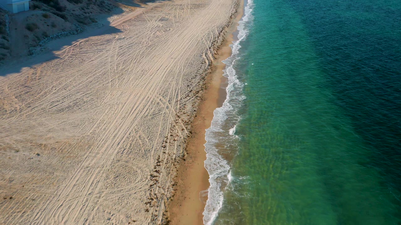 Top-down coastal view of sand tracks, surf, and green water