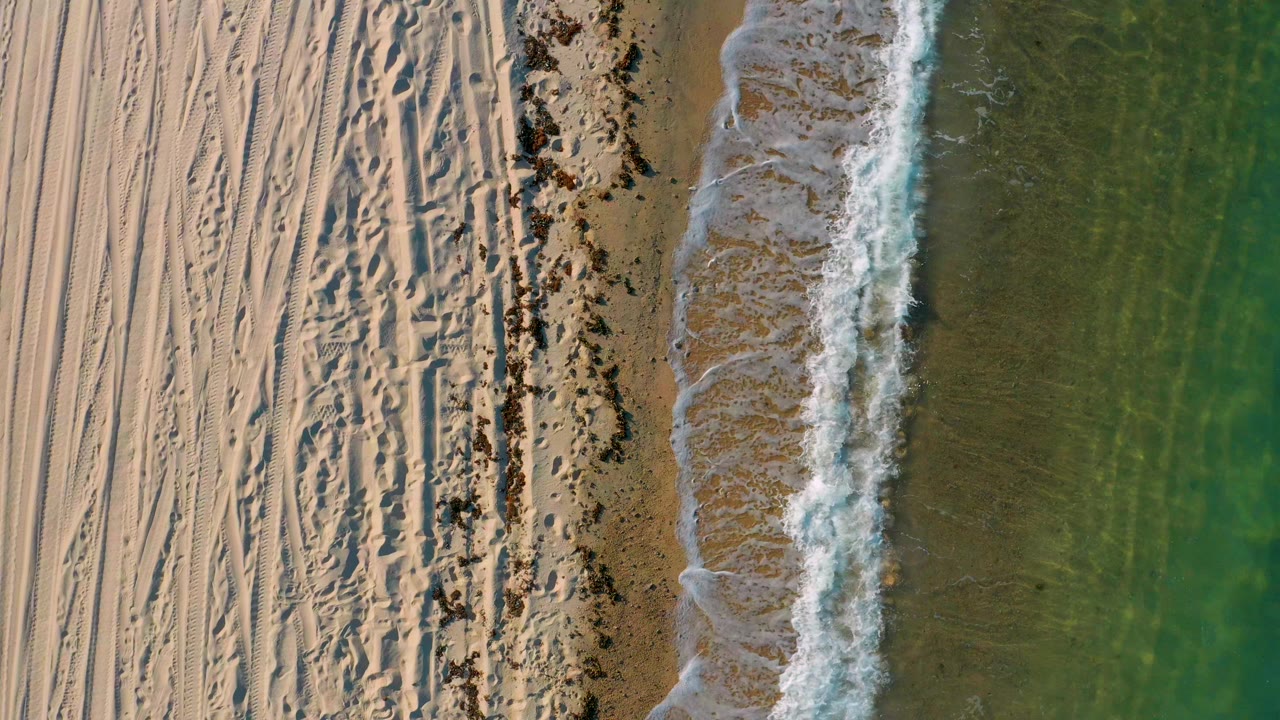 Overhead view in El Sargento La Ventana, Mexico, with pale tracked sand on the left, a dark shoreline strip, white surf at center, green sea on the right, and no visible people.