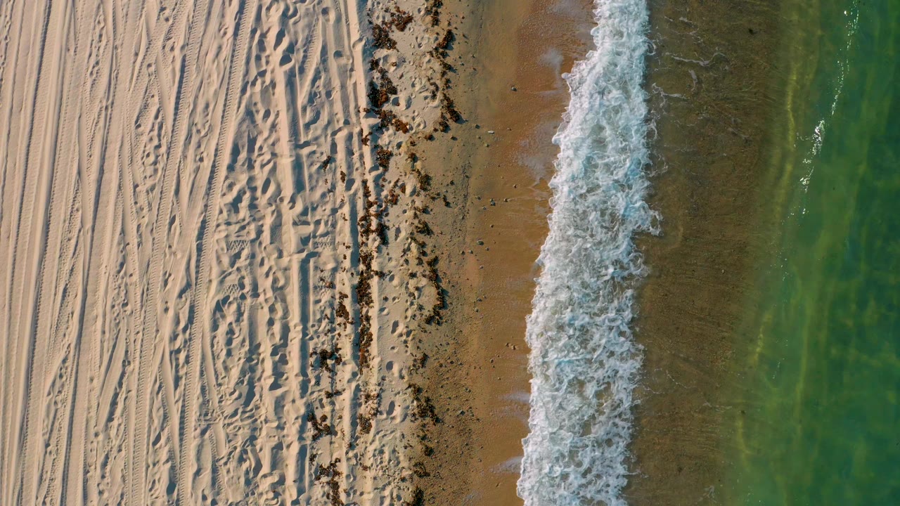 In El Sargento La Ventana, Mexico, a bright white breaker runs down the shoreline, separating tan sand and a dark wrack line from green water, with no visible people.