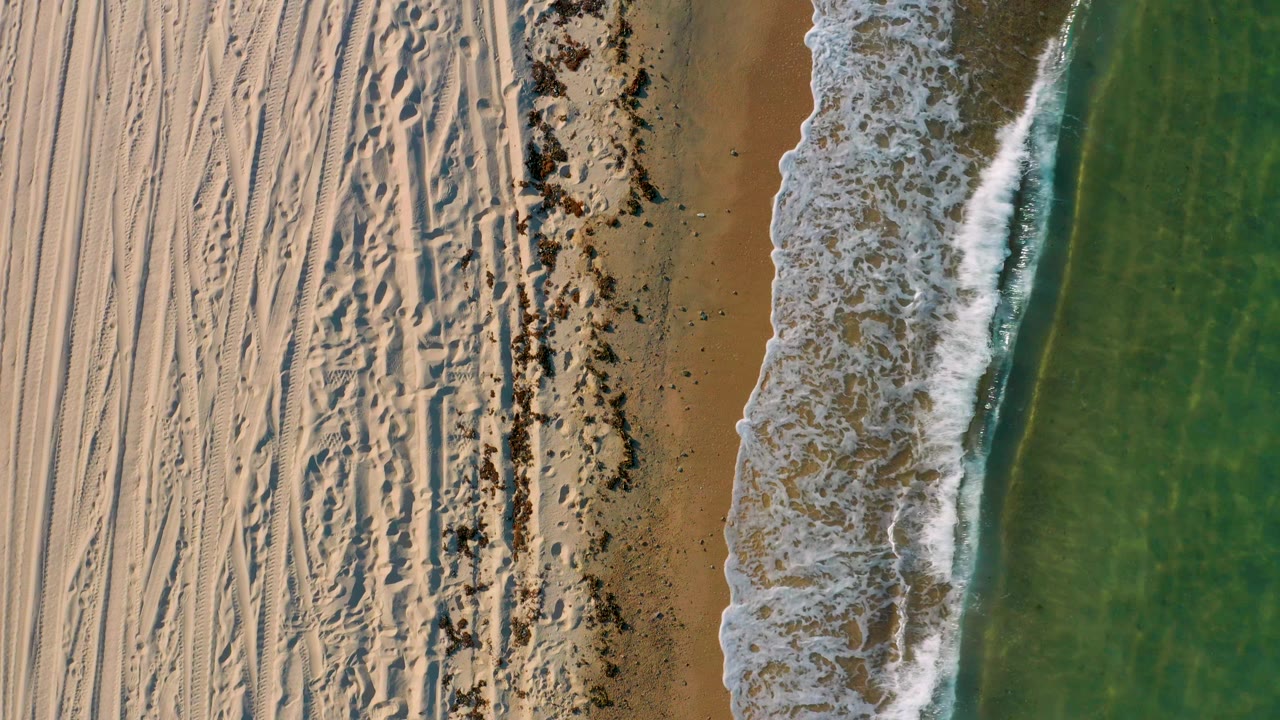 The shoreline at El Sargento La Ventana appears from above with rippled and tracked sand, scattered dark beach material, a foamy breaking wave, and green-blue water, no visible people.