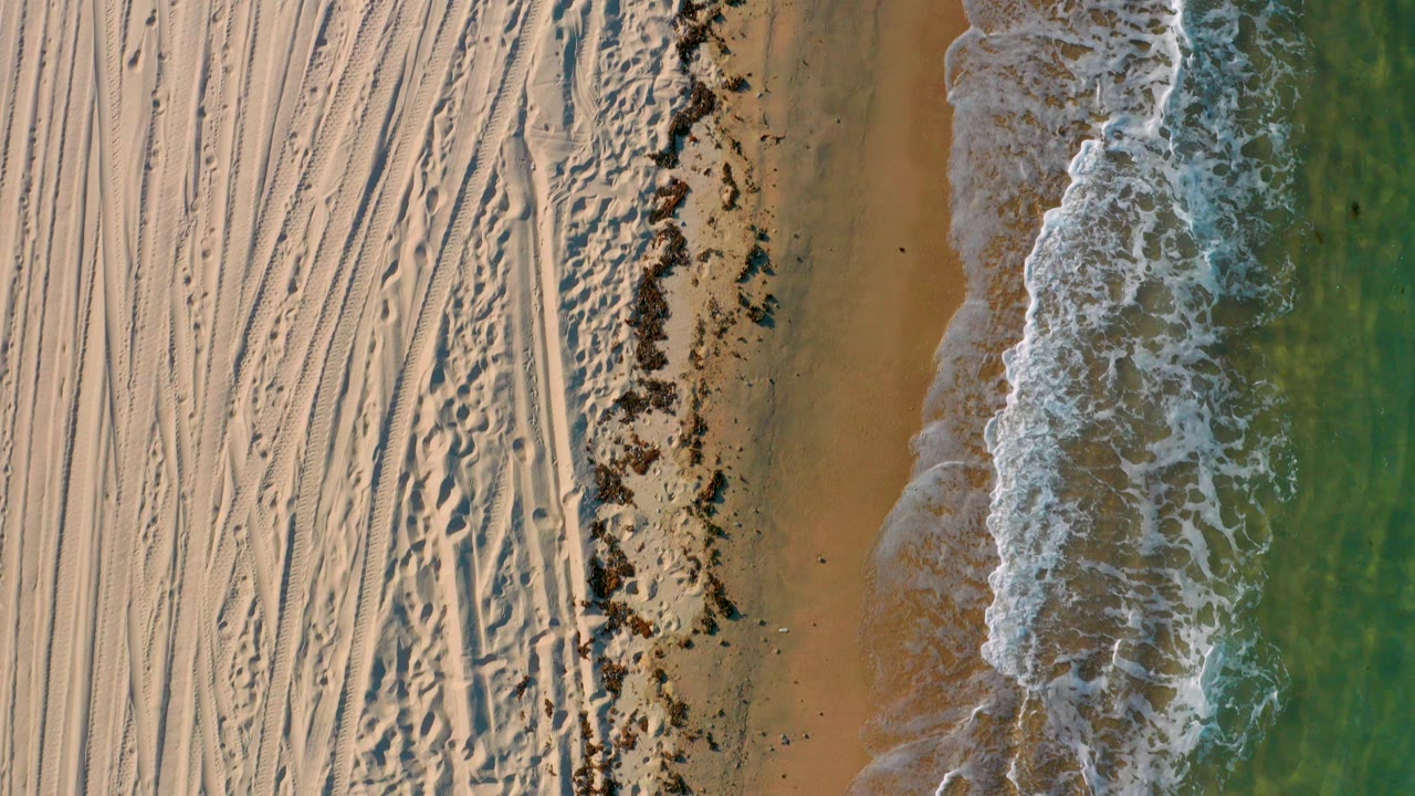 Overhead view in El Sargento La Ventana, Mexico, with tracked beige sand at left, wet shoreline through the center, green water at right, and no visible people.