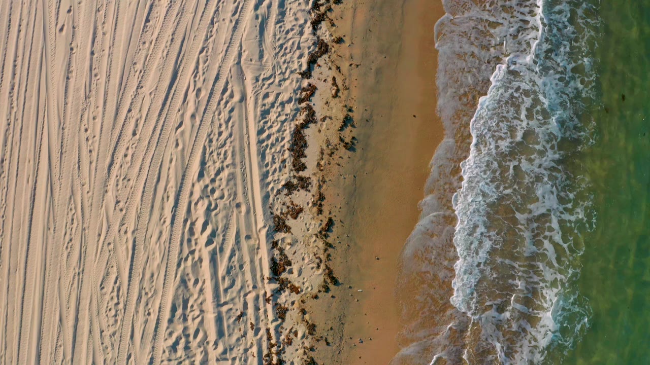 Aerial frame of the Mexico shoreline with the foamy tide slightly farther along the beach edge, tracked sand on the left, green water on the right, and no visible people.