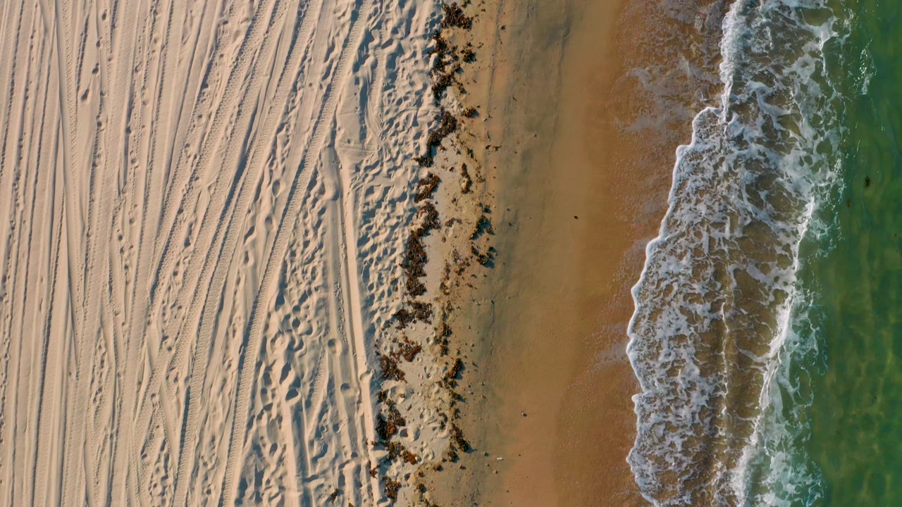 Top-down view in El Sargento La Ventana, Mexico, showing a larger curl of white foam over the wet beach, textured sand tracks on the left, green water on the right, and no visible people.