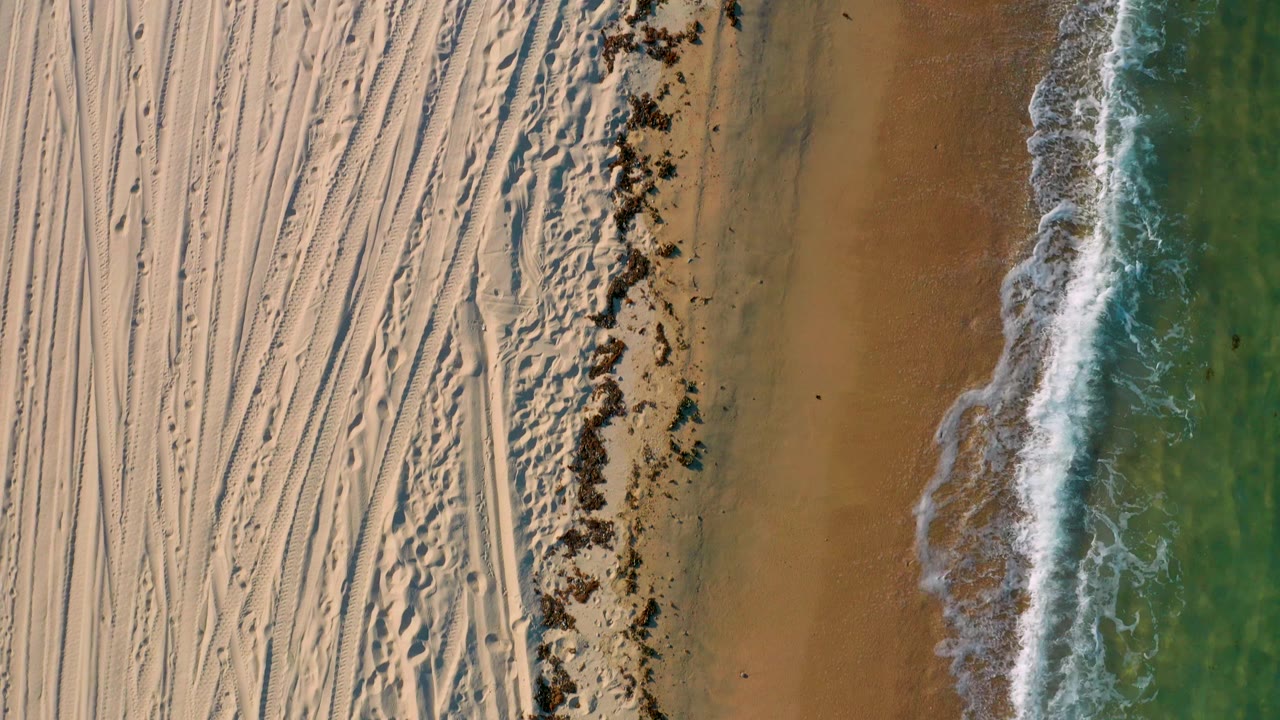 Overhead El Sargento La Ventana shoreline with dense tire and foot tracks in dry sand, a darker uneven beach band, thin white surf, green ocean, and no visible people.
