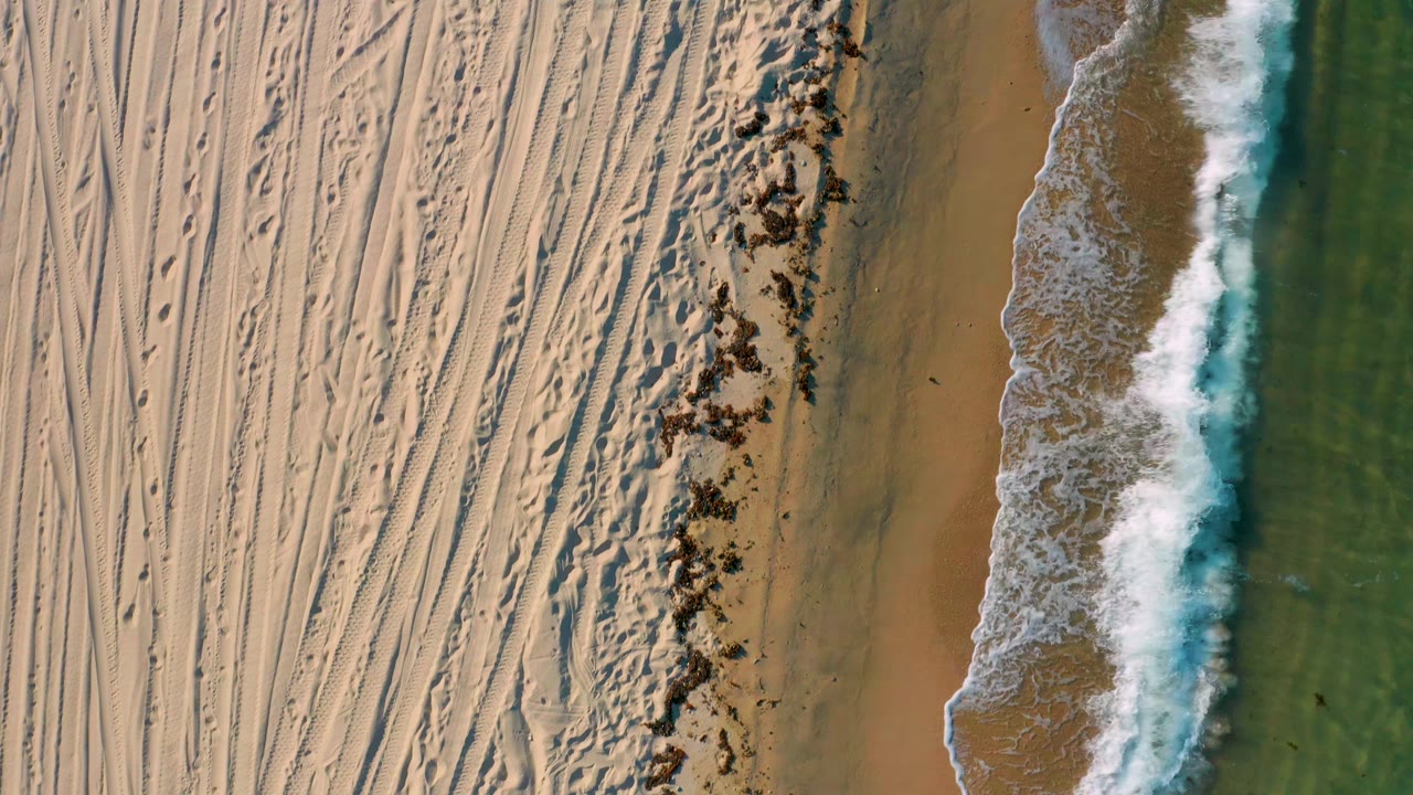 Aerial frame from El Sargento La Ventana, Mexico, with dense tire-like tracks across dry sand, a darker wrack or rock strip, and a breaking wave pressed close to shore with no visible people.