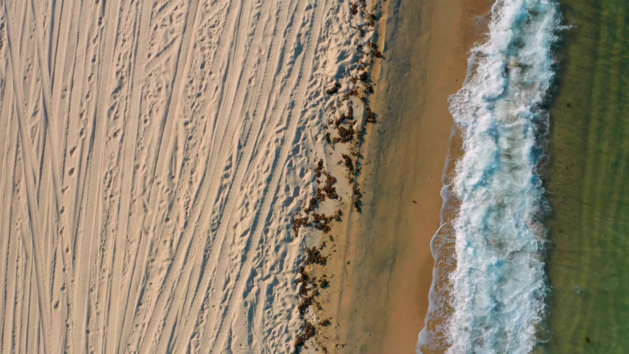 Top-down El Sargento La Ventana shoreline frame showing pale sand cut by parallel tracks, brown wet sand, green water, and a wider white foam edge, with no visible people.