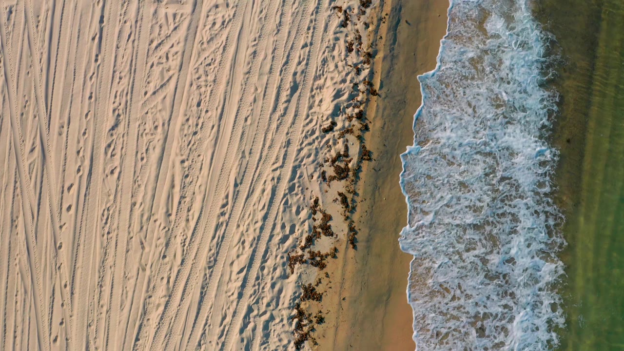 Overhead frame in El Sargento La Ventana, Mexico, where a larger sheet of white wave wash covers the wet sand beside the tracked dry beach, with no visible people.