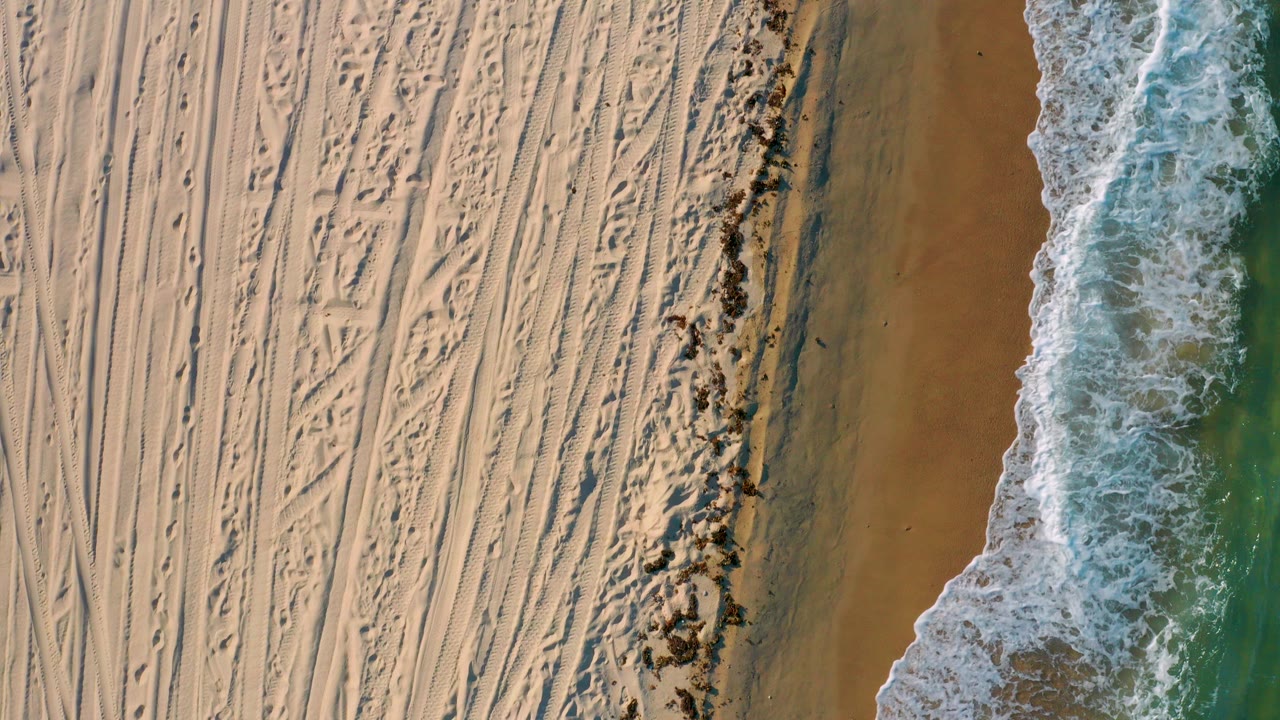 Top-down aerial view of El Sargento La Ventana beach, with pale sand tracks on the left, a dark seaweed line near center, green-blue surf on the right, and no visible people.