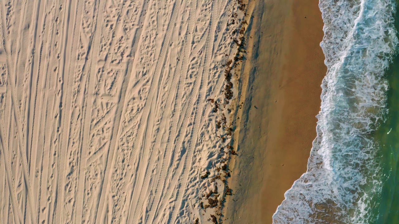 Aerial beach view with parallel sand tracks beside breaking waves.