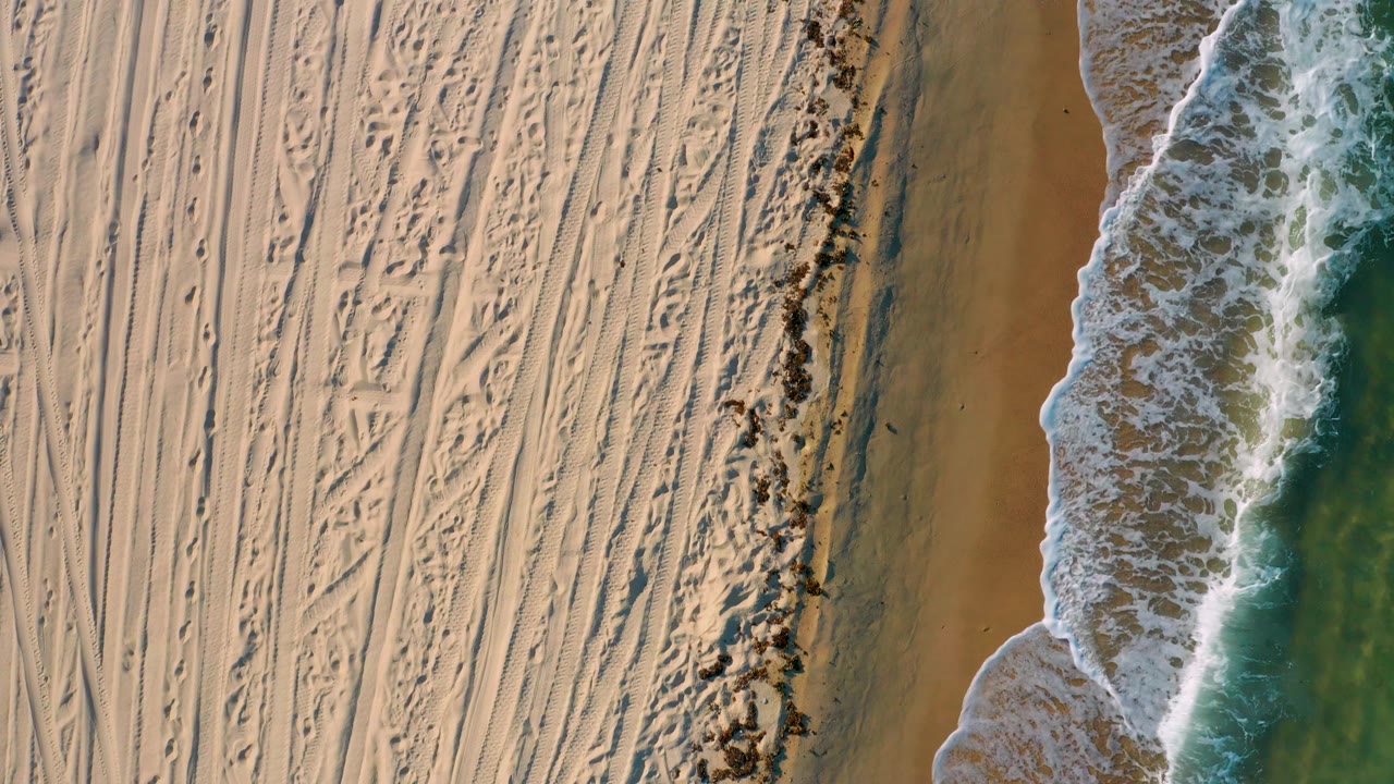 Overhead view of El Sargento La Ventana shoreline, where tracked sand fills the left of frame and a curving edge of white surf meets darker wet sand on the right, with no visible people.