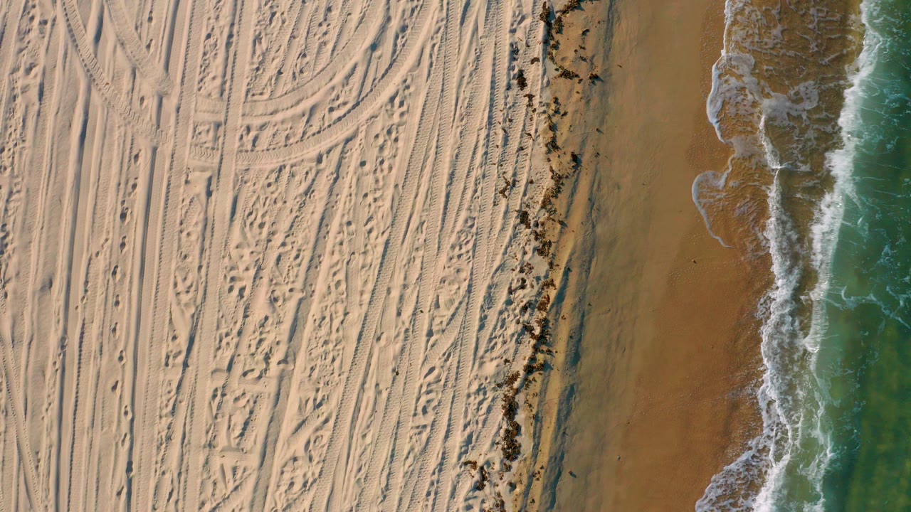 Straight-down view of El Sargento La Ventana beach with tracked pale sand on the left, a narrow debris line, and green-blue surf breaking on the right, with tiny people faintly visible.