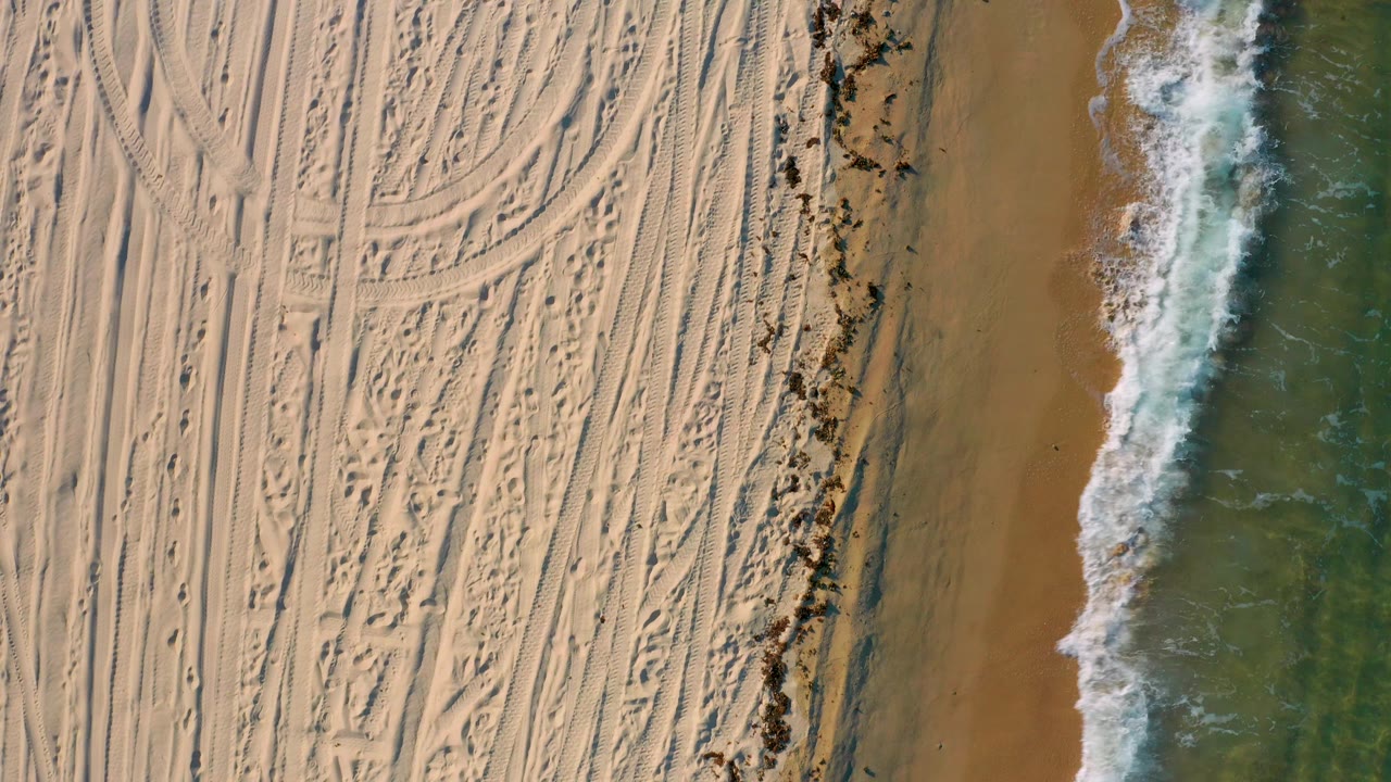 Straight-down aerial view of El Sargento La Ventana with pale tracked sand filling most of the frame and white surf advancing along the right side over green water.