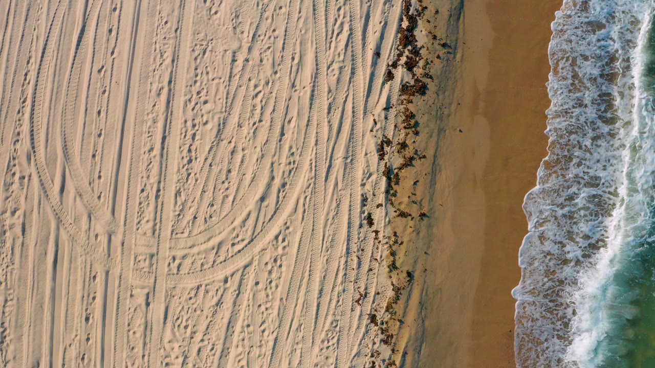 Top-down view in El Sargento La Ventana, Mexico, showing pale sand crossed by tire marks with a narrow band of foamy surf along the right edge and no visible people.