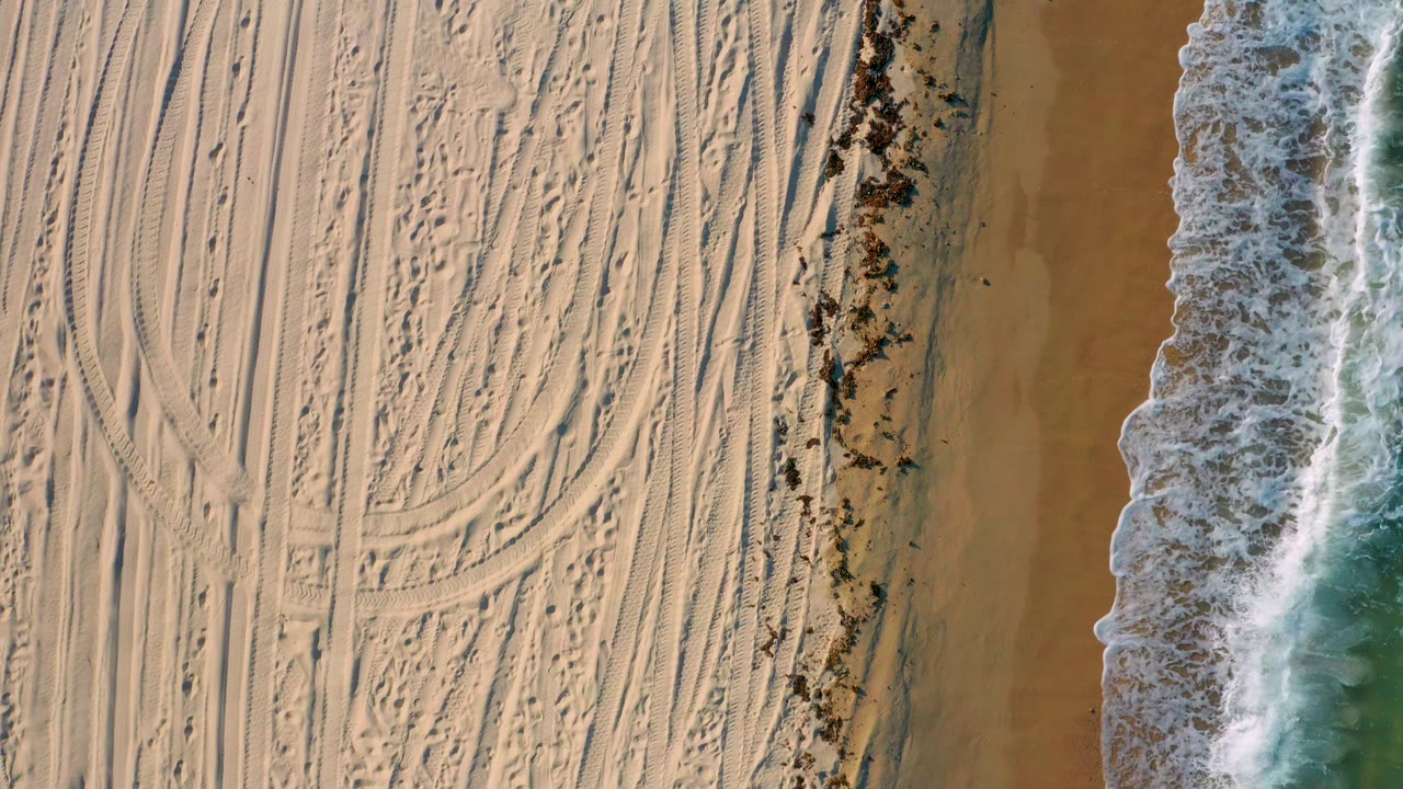 Aerial frame in El Sargento La Ventana, Mexico, with dense curved tire marks across the left sand and white surf advancing along the right side, with no visible people.