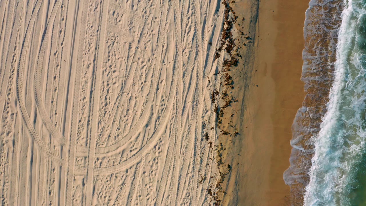 Overhead view in El Sargento La Ventana, Mexico, where parallel and arcing tire tracks fill the sand while green-blue water and foam line the frame edge, with no visible people.