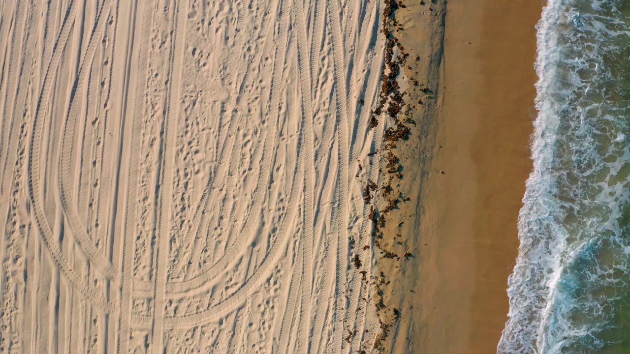 Top-down El Sargento La Ventana, Mexico beach view with dry sand patterned by tire marks, a dark wrack line near the wet sand, and a thin foamy wave edge, with no visible people.