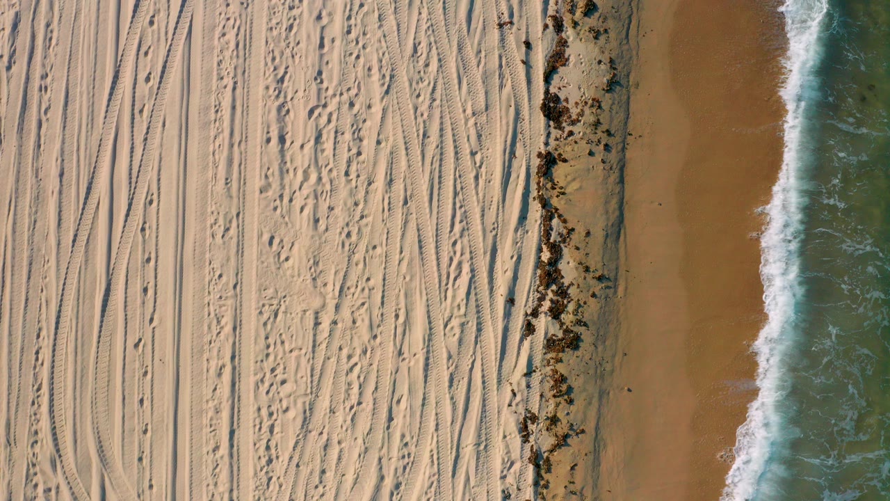 Overhead view in El Sargento La Ventana, Mexico showing tracked pale sand, a dark seaweed band, and green-blue surf along the right edge, with no visible people.