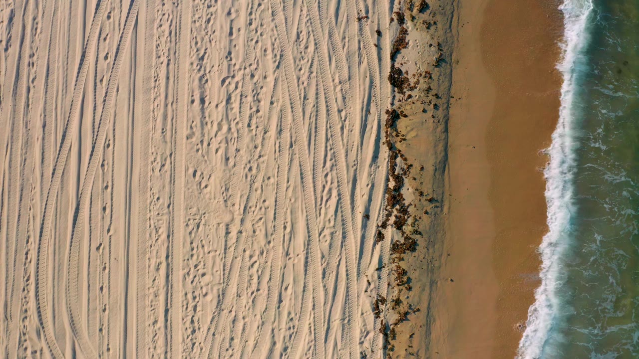 Overhead frame from El Sargento La Ventana, Mexico with dense sand tracks on the left, a dark seaweed strip near the shore, and a narrow white wave line on the right, with no visible people.