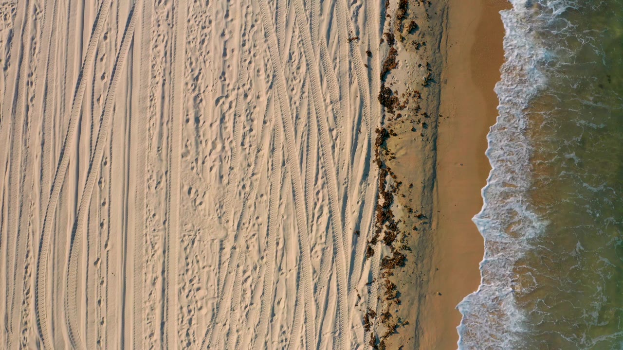 Aerial view in El Sargento La Ventana, Mexico where the surf spreads farther across the wet brown sand beside the dark seaweed line, with no visible people.