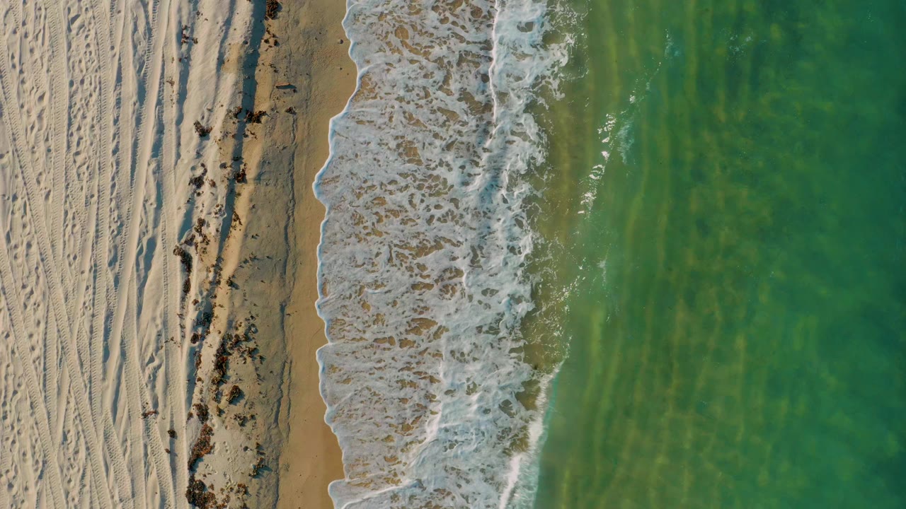 Overhead view of El Sargento La Ventana shoreline with pale sand at left, a foamy wave band near center, green-blue water at right, and no visible people.