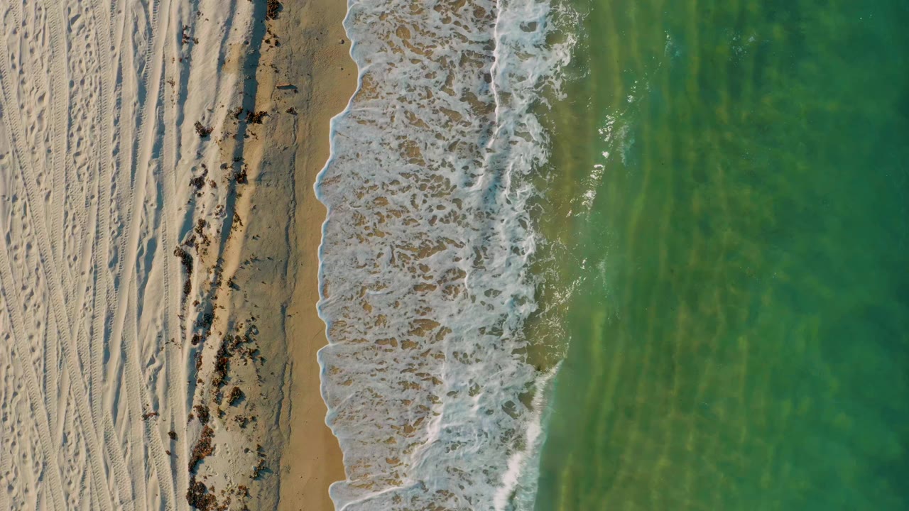 Aerial frame from El Sargento La Ventana shows the surf line slightly farther along the sand, with textured beach on the left, translucent green water on the right, and no visible people.