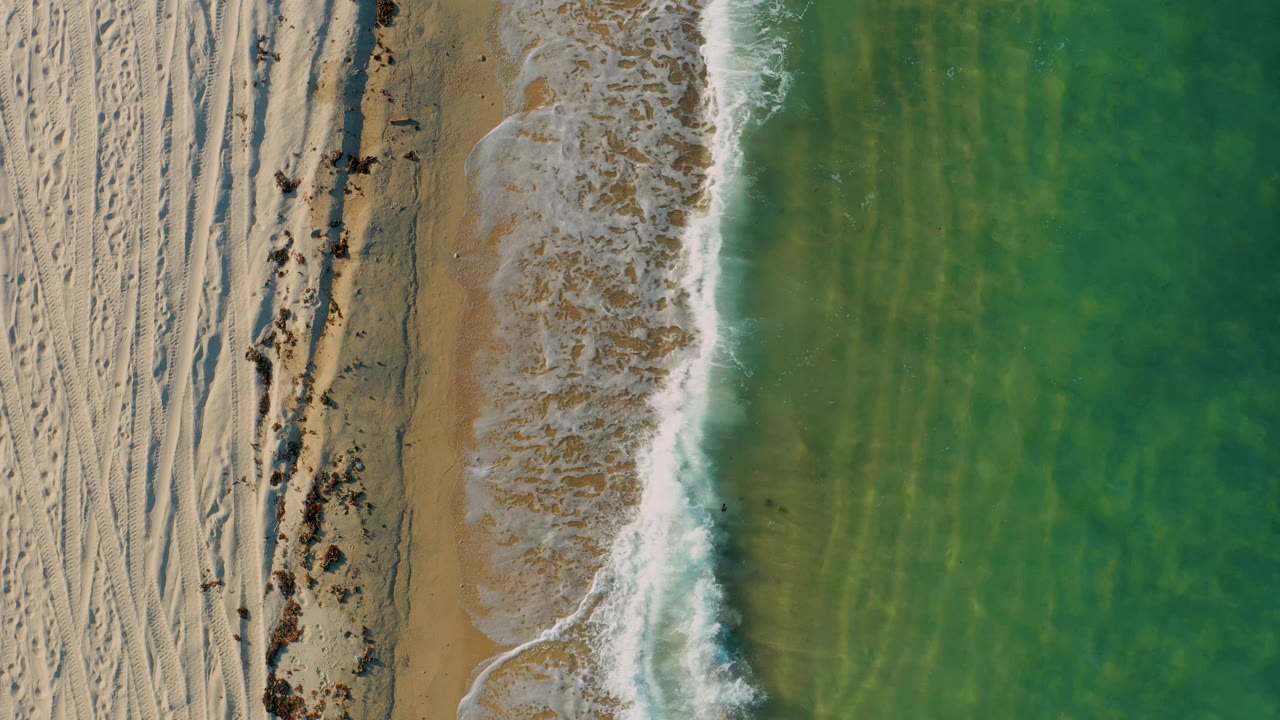 Top-down view in El Sargento La Ventana with a broader patch of white foam washing over tan sand, clear green water filling the right side, and no visible people.