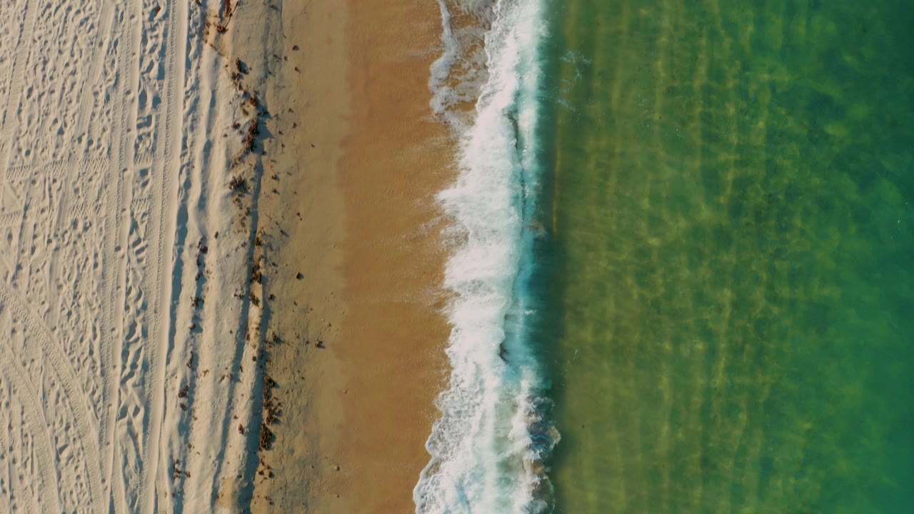 Overhead view of El Sargento La Ventana shoreline with pale dry sand on the left, a narrow foam edge, clear green water on the right, and no visible people.