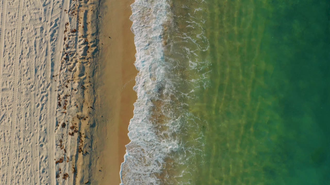 Overhead frame from El Sargento La Ventana showing a small wave breaking diagonally along the sand with clear shallow water to the right and no visible people.