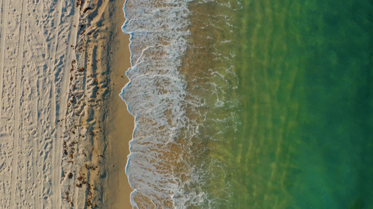 Top-down El Sargento La Ventana beach view with rippled sand at left, a thicker band of foamy surf near the center, clear green water at right, and no visible people.