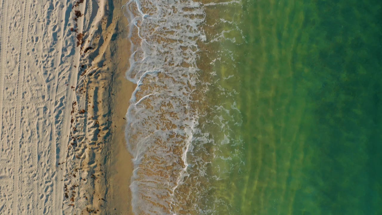 Overhead view in El Sargento La Ventana where foamy water spreads across tan wet sand, with green-blue shallows occupying the right side and no visible people.