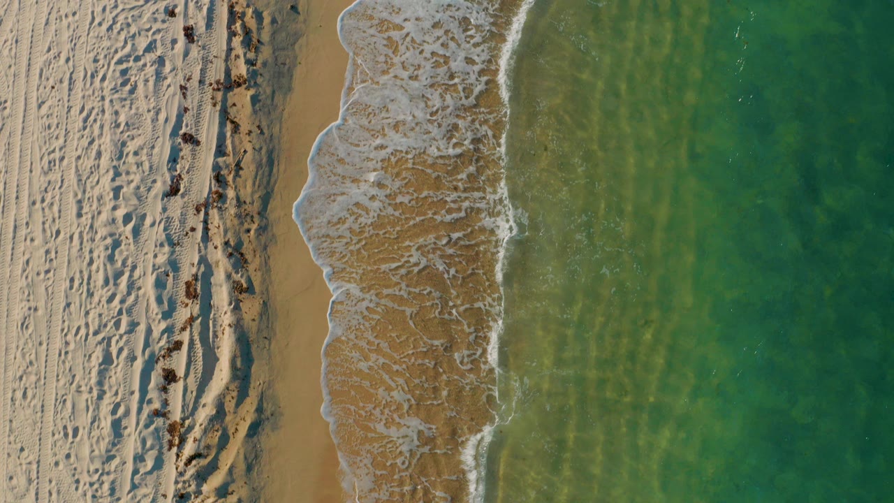 Top-down view of El Sargento La Ventana shoreline with pale sand at left, white foam at the waterline, green-blue sea at right, and no visible people.