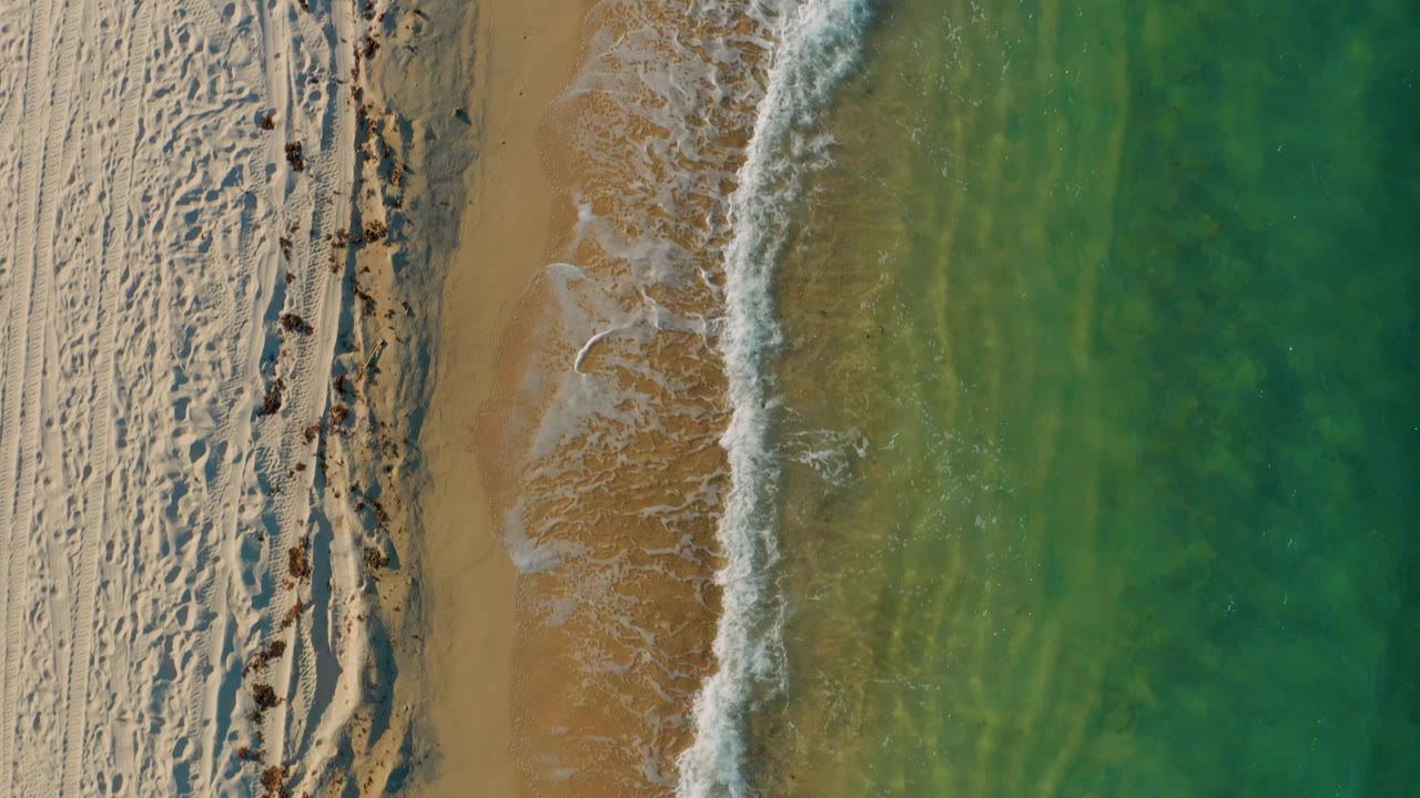 Overhead view of El Sargento La Ventana shoreline with textured sand, a brighter surf band, and broad turquoise-green water filling the right side, with no visible people.