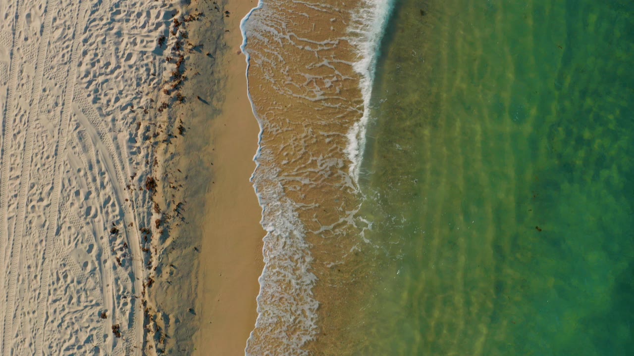 Overhead view of El Sargento La Ventana shoreline with pale textured sand on the left, a narrow surf line in the center, green shallow water on the right, and no clearly visible people.