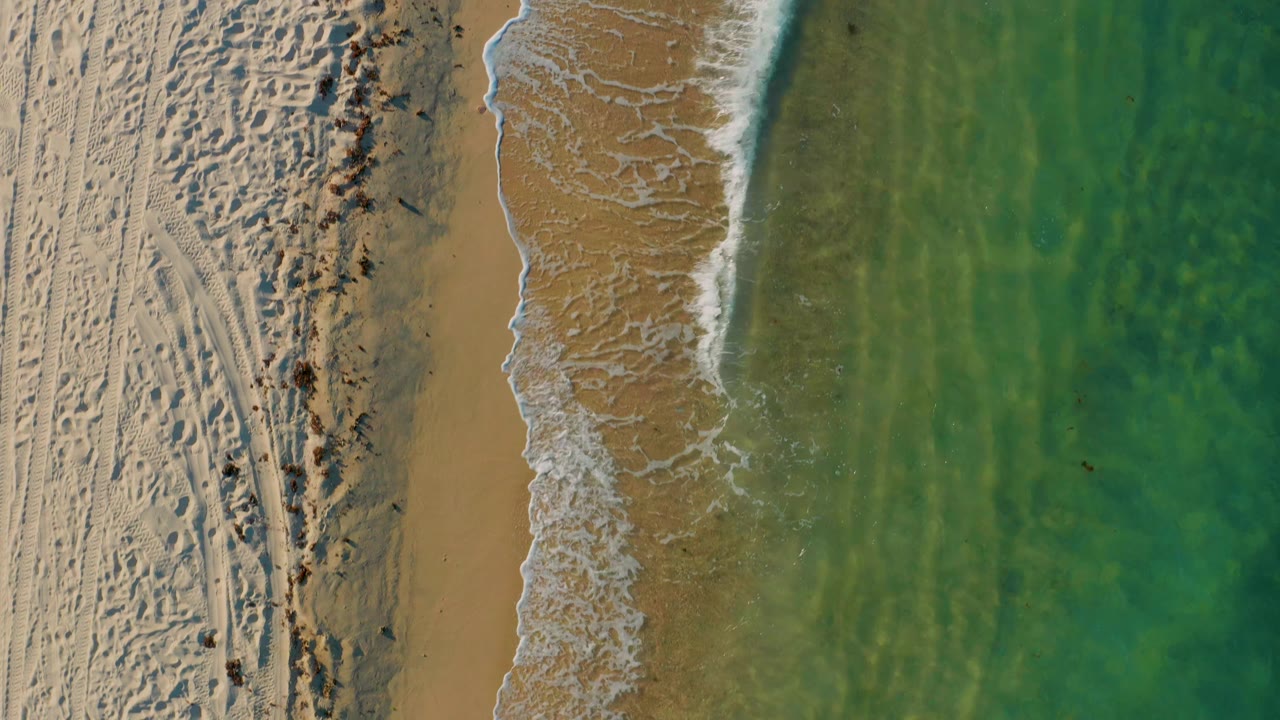 Aerial frame over El Sargento La Ventana showing a straighter band of white foam along wet sand, with rippled green water filling the right side and no clearly visible people.