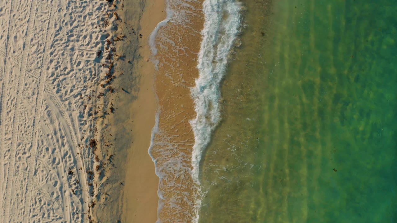 Top-down El Sargento La Ventana beach view with bright sand texture, a thicker breaking wave near the shore, green water to the right, and no clearly visible people.