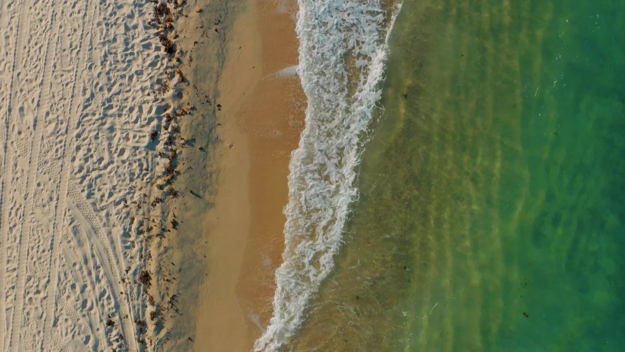 Overhead view of El Sargento La Ventana with rough sand texture and tracks on the left, a foamy surf strip through the center, green water on the right, and no clearly visible people.