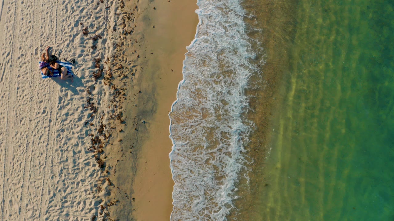Overhead view in El Sargento La Ventana, Mexico, with green water on the right, white surf along wet sand, textured beach tracks on the left, and small distant beachgoers near the upper left.