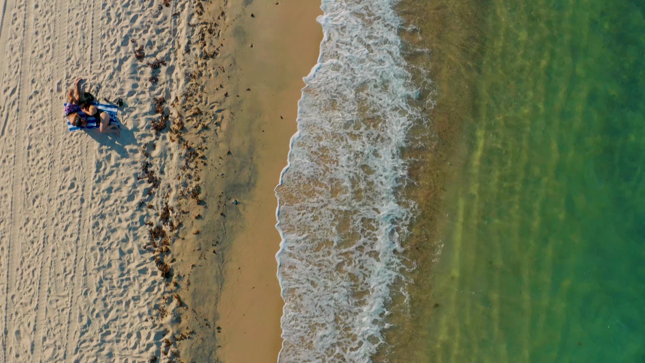 Aerial frame from El Sargento La Ventana, Mexico, showing a vertical band of foamy shore break between tan sand and green ocean, with small distant beachgoers still visible on the upper left beach.