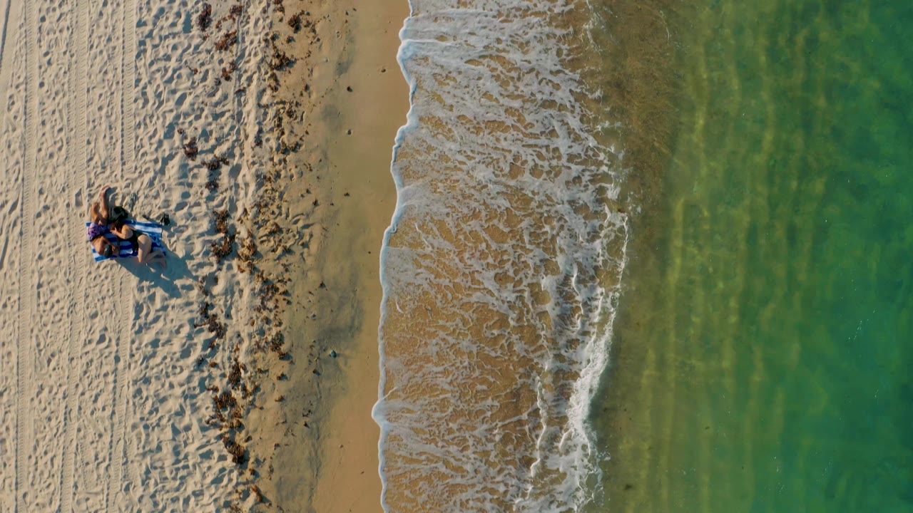 Top-down view in El Sargento La Ventana, Mexico, with the surf curling along a narrow strip of wet sand, green water filling the right half, beach tracks at left, and small distant beachgoers near the left side.