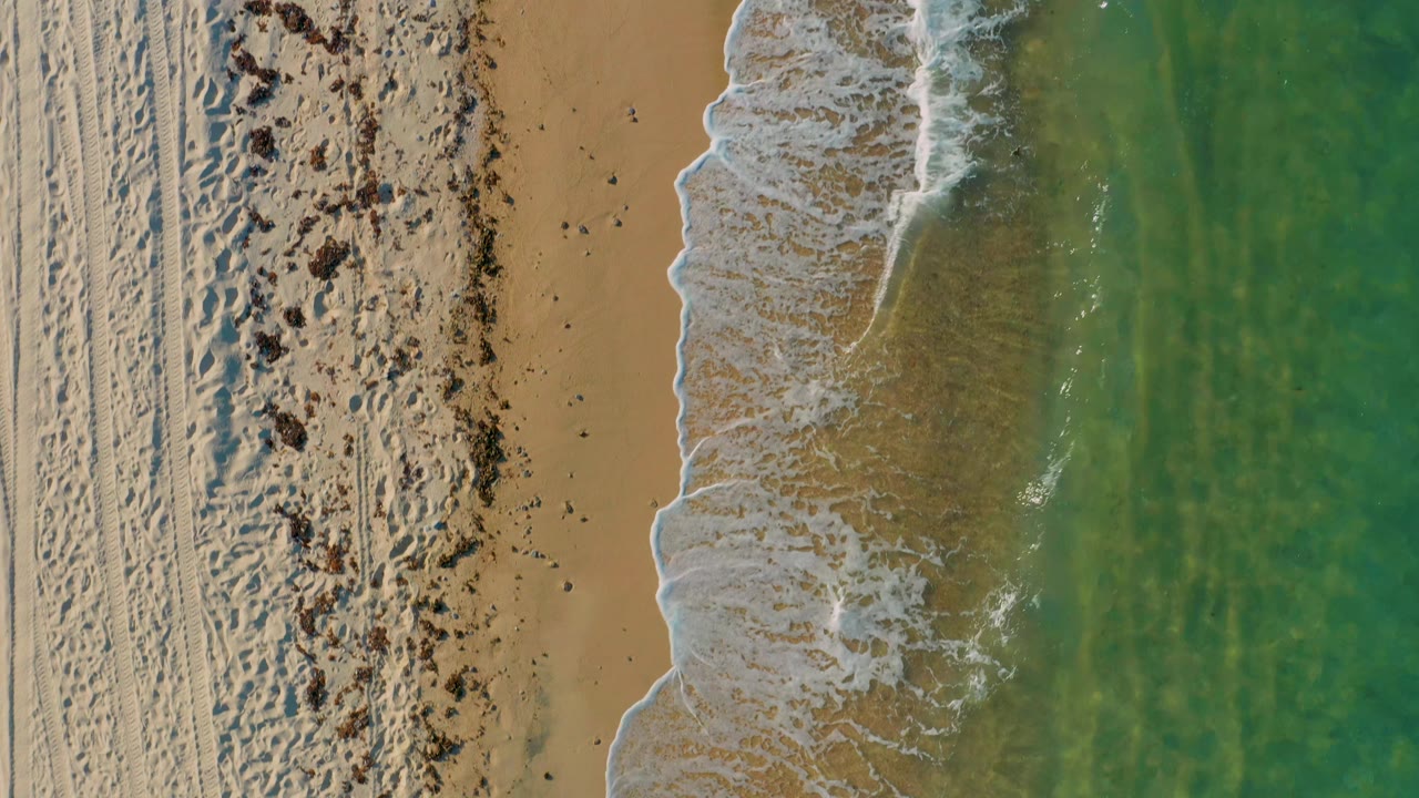 A near-vertical aerial frame at El Sargento La Ventana shows a curved band of white foam pushing onto tan sand with green ocean water to the right and no visible people.