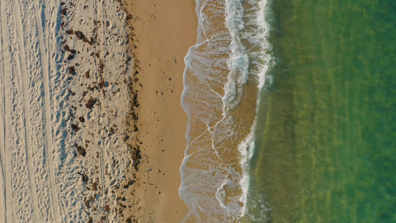 Overhead El Sargento La Ventana beach view with parallel tire tracks on pale sand, dark seaweed or debris patches, a foamy shore break, green water, and no visible people.