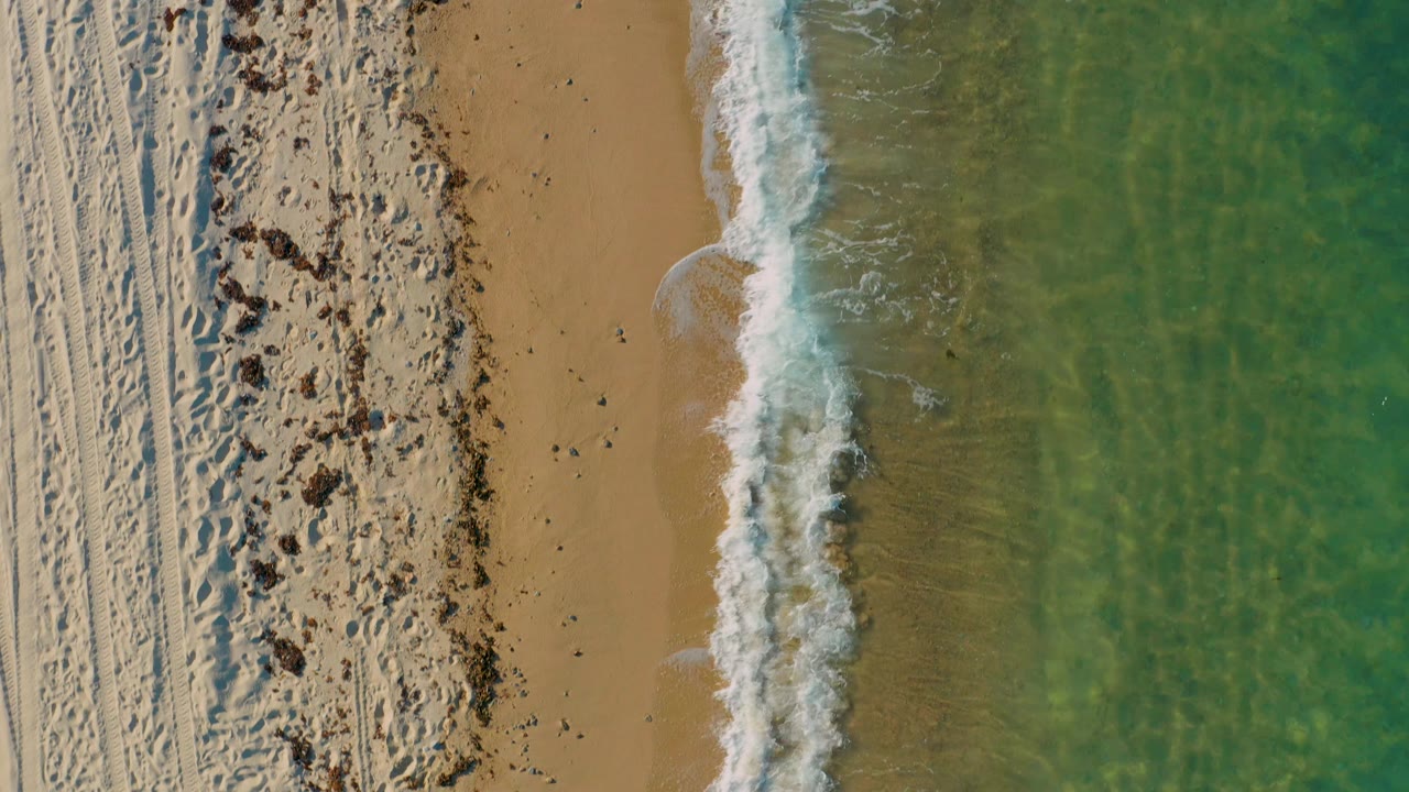 Aerial shoreline frame from El Sargento La Ventana with a vertical strip of breaking white surf, open tan sand on the left, green shallow water on the right, and no visible people.