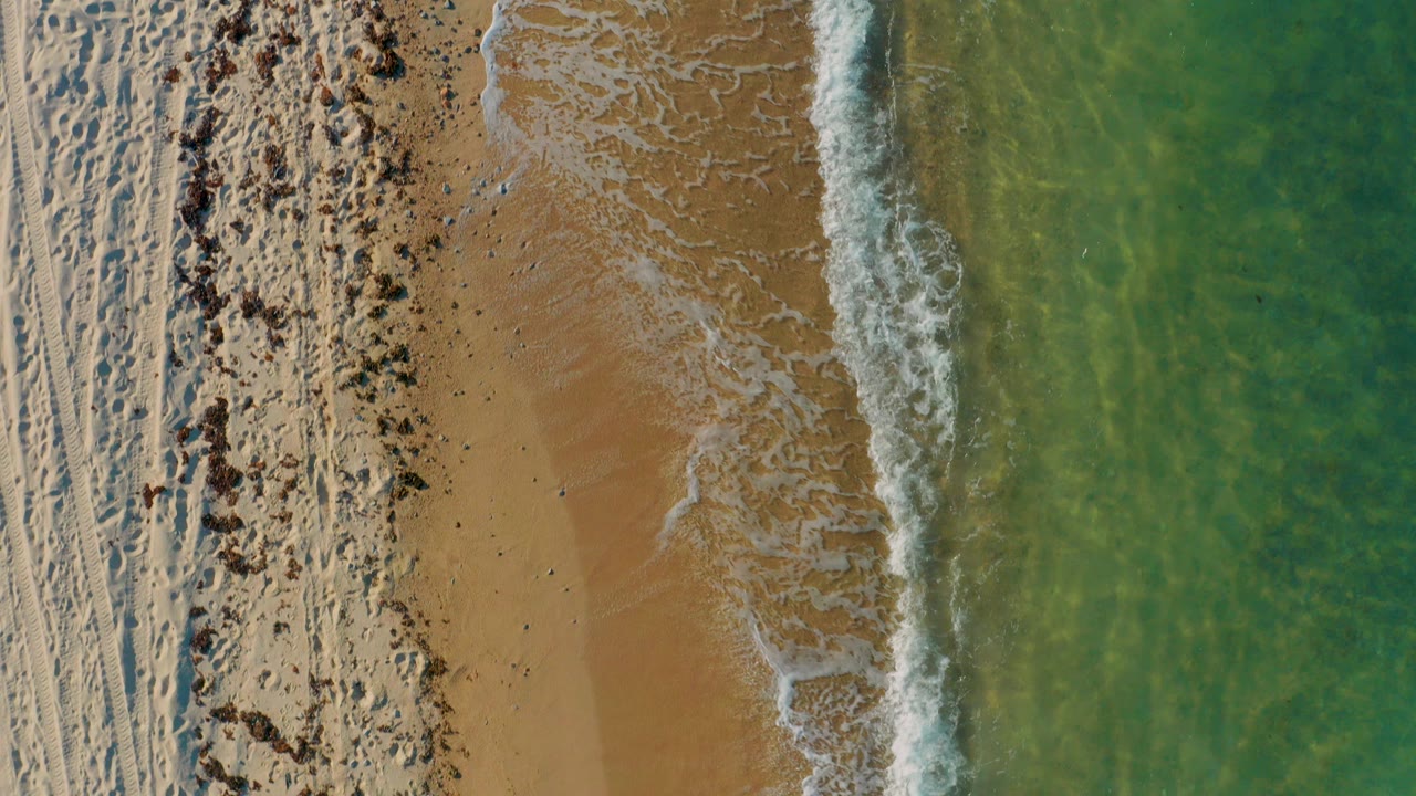 Top-down aerial view of El Sargento La Ventana shoreline with pale beach tracks on the left, tan wet sand near the center, clear green-blue water on the right, and no visible people.