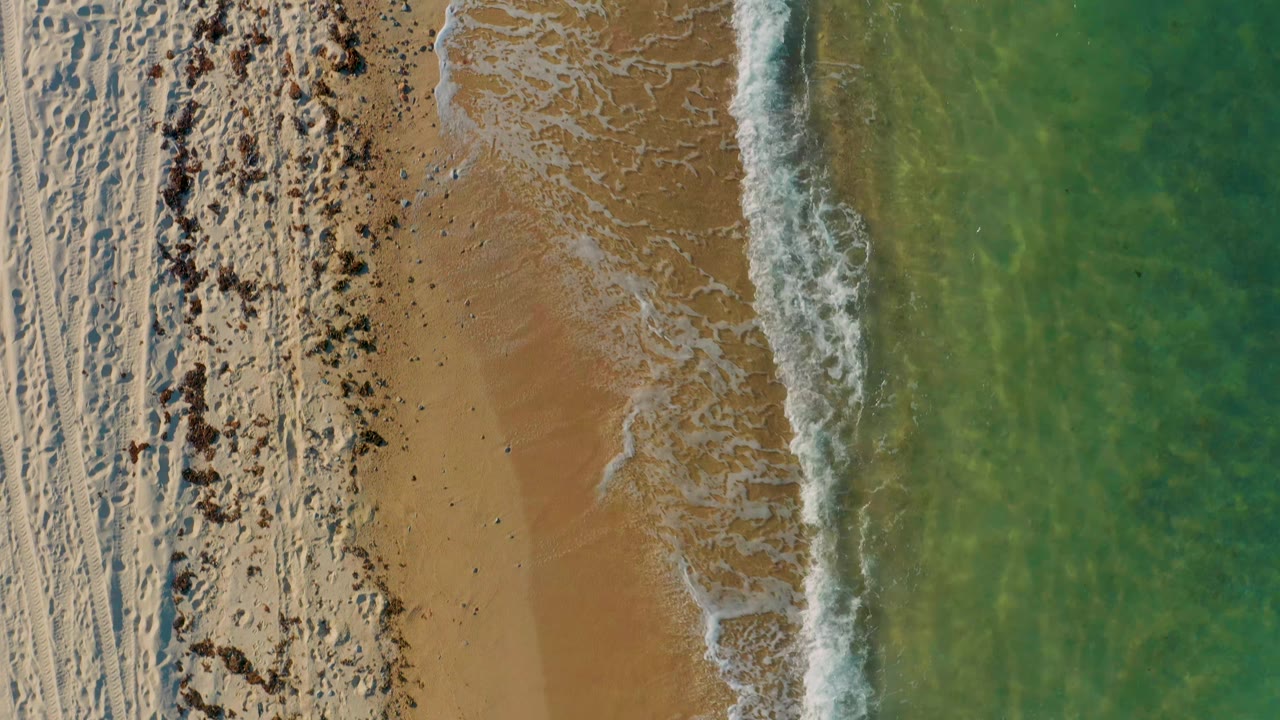 Overhead frame from El Sargento La Ventana, Mexico, showing a narrow band of white foam curling onto tan sand, textured pale beach at left, green water at right, and no visible people.