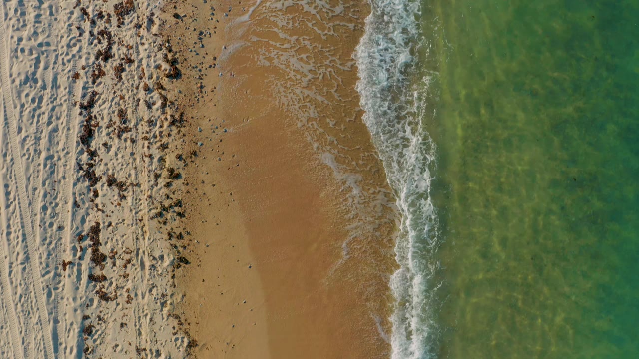 Aerial view over the El Sargento La Ventana coast with a curved white surf line, open tan sand, rougher pale beach texture to the left, clear turquoise water to the right, and no visible people.